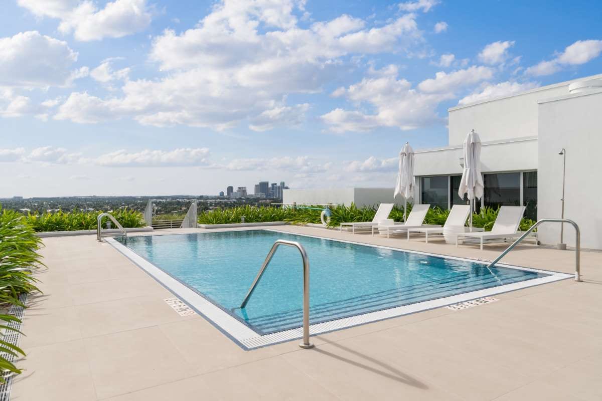 Modern building beside a pool with trees, chairs, and blue sky reflected in water