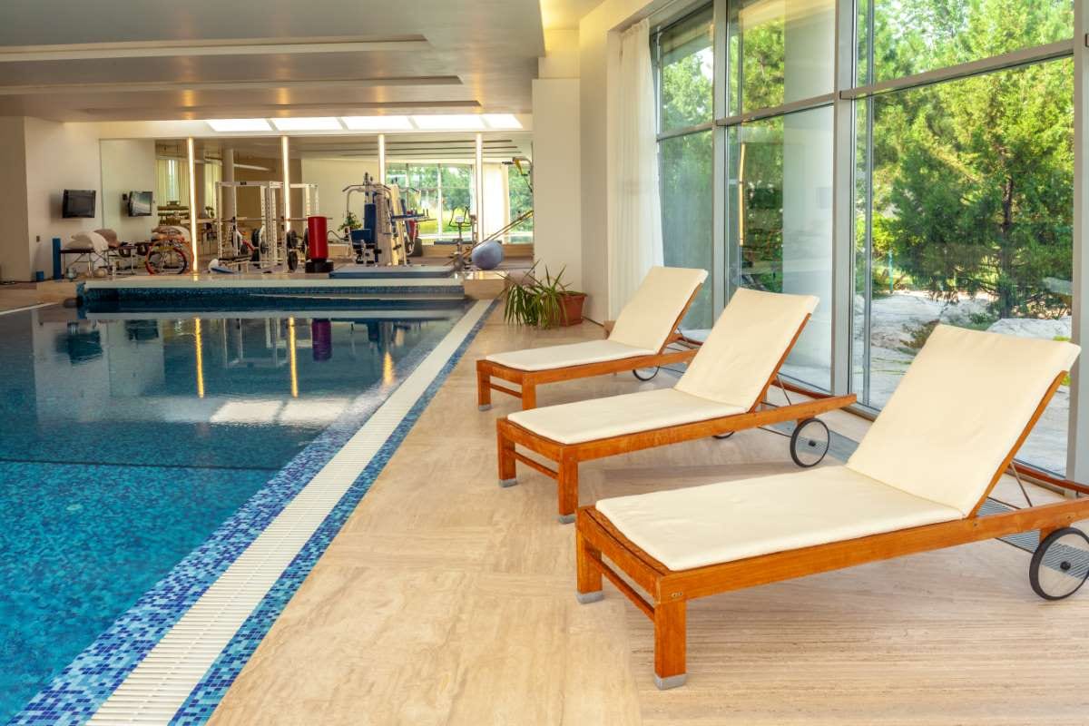 Wooden chaise lounges beside an indoor pool with conifer trees outside the window